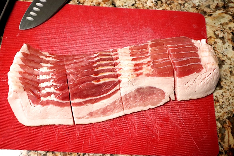 traditional bacon cut into quarters lengthwise on top of a red cutting board with a knife in the background