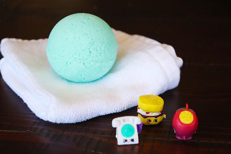 A blue bath bomb filled with a toy, setting on top of a white washcloth with 3 shopkins toys in front 